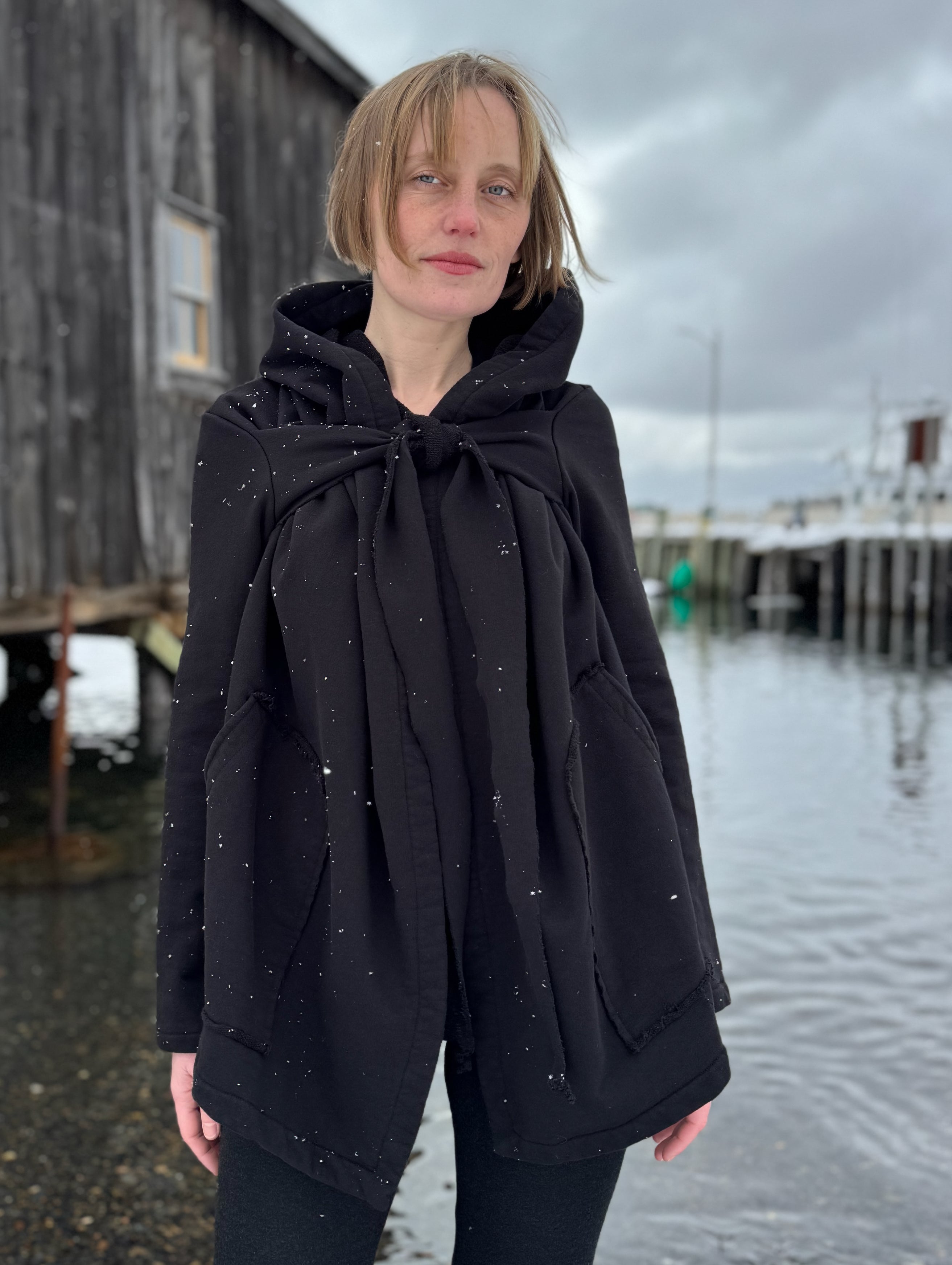 Person wearing a black hooded coat standing by a waterfront on a cloudy day.