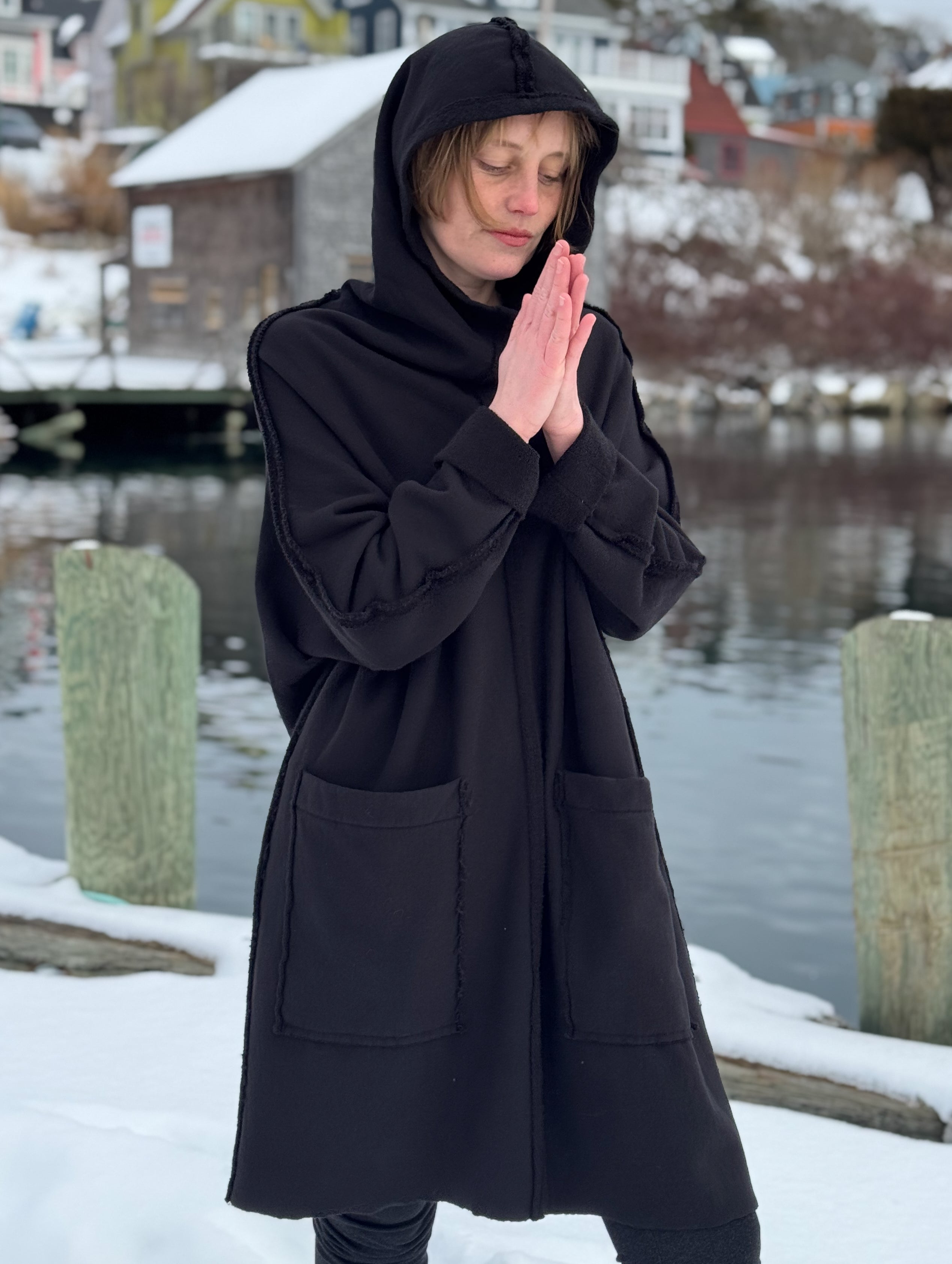 Person wearing a black hooded coat in a snowy landscape with water and buildings in the background.