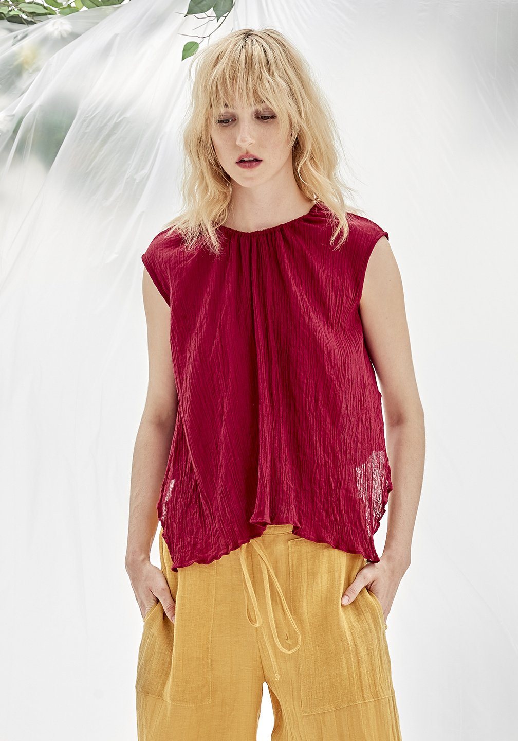 A sleeveless red organic cotton crepe top with a gathered neckline, paired with yellow pants. The person is standing against a white background with a floral pattern.