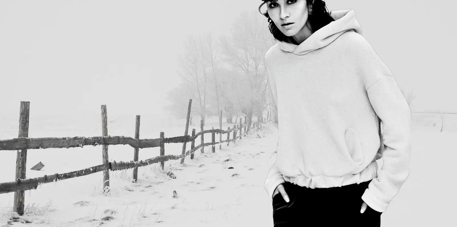 Person wearing a light-colored hoodie standing in a snowy landscape with a wooden fence.
