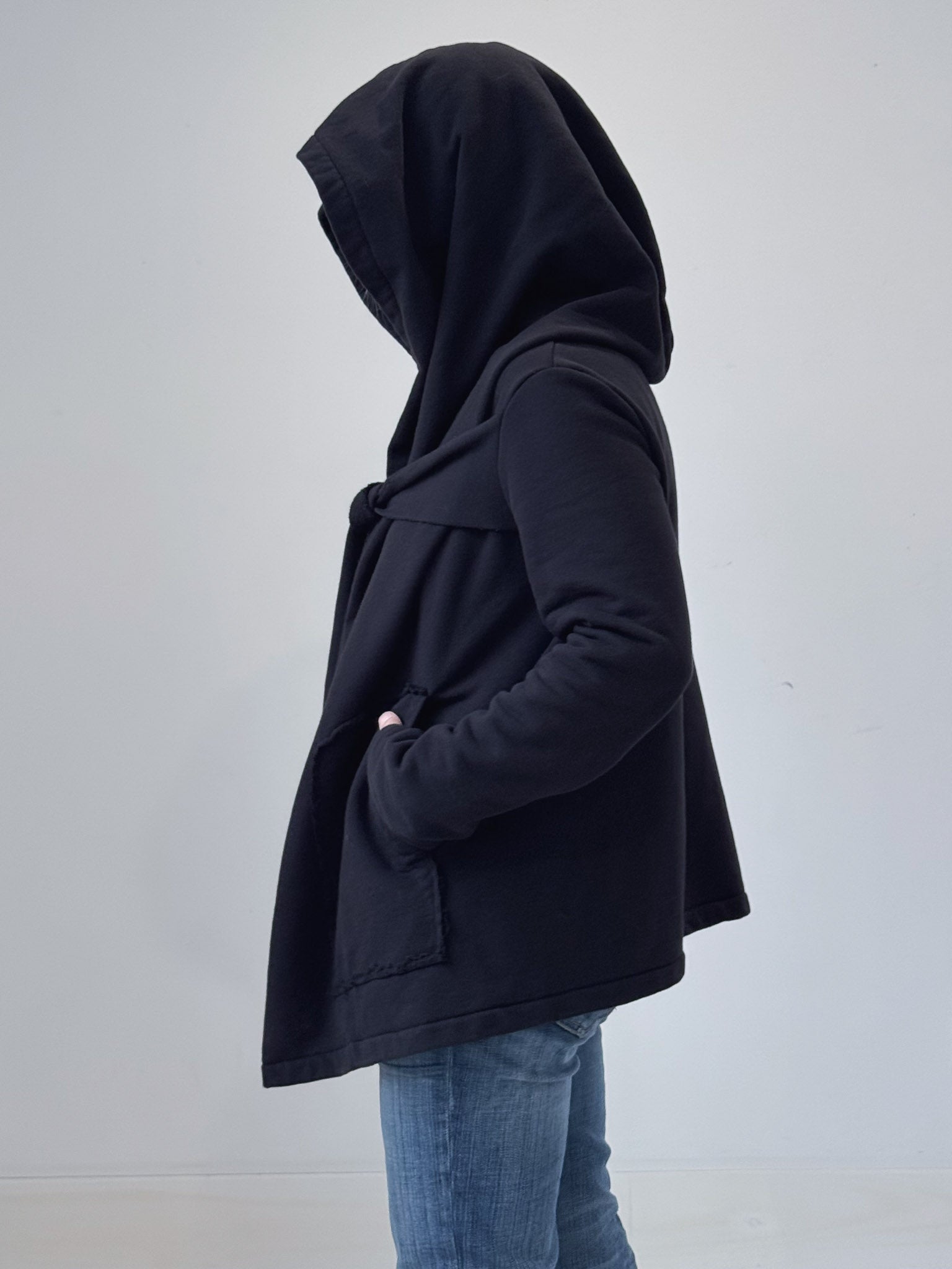 Person wearing a black hooded jacket and blue jeans on a plain background