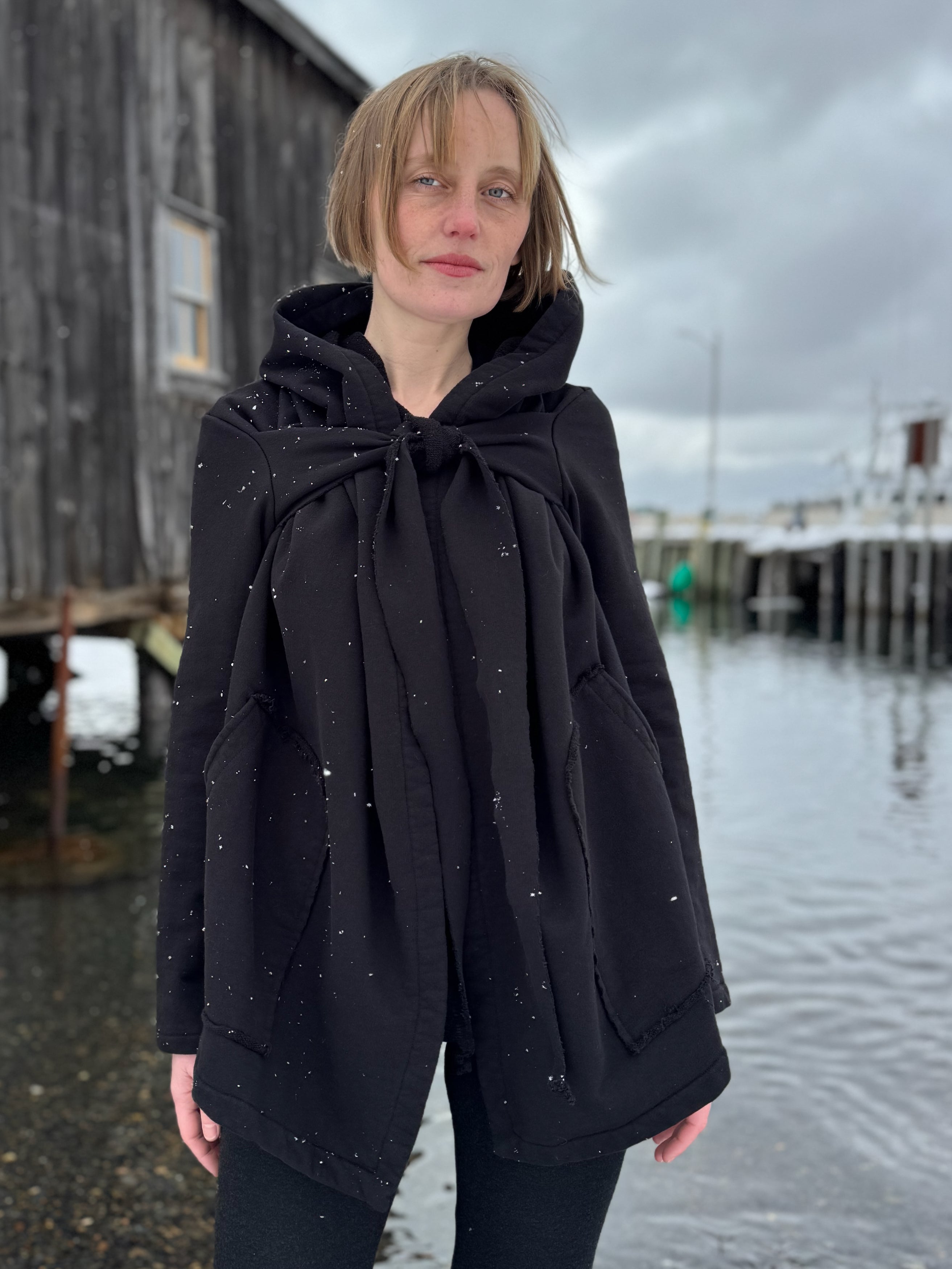 Person wearing a black hooded coat standing by a waterfront on a cloudy day.