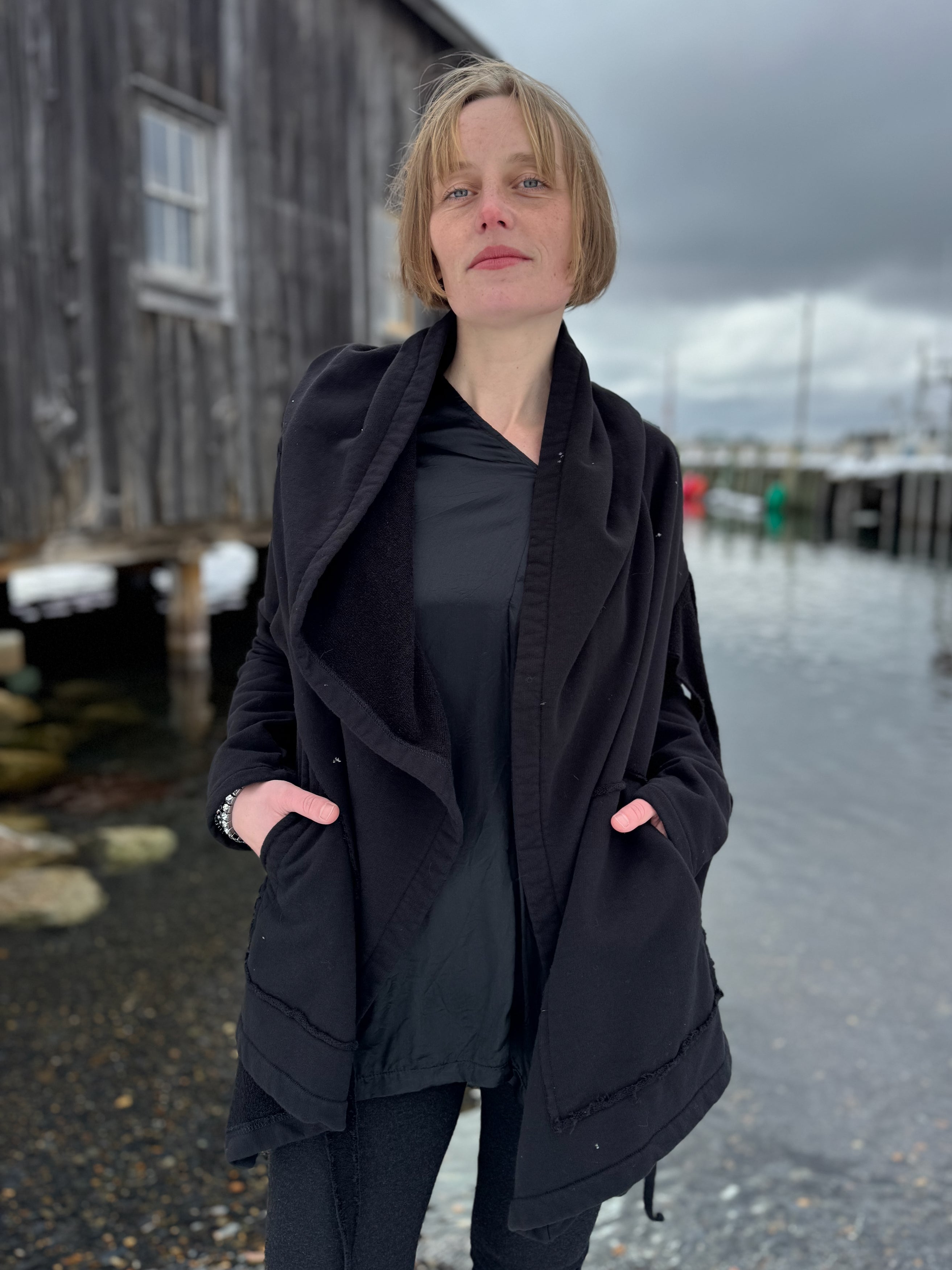 Person wearing a black coat standing outdoors with a wooden building and cloudy sky in the background