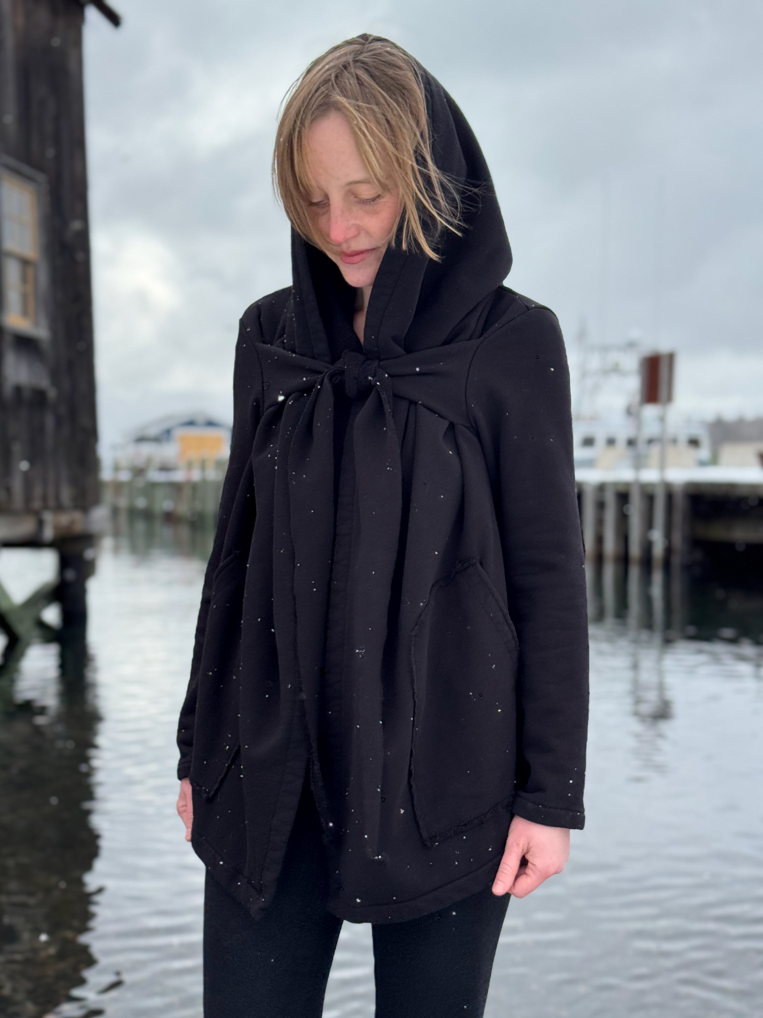 Person wearing a black hooded coat standing on a dock with a cloudy sky.