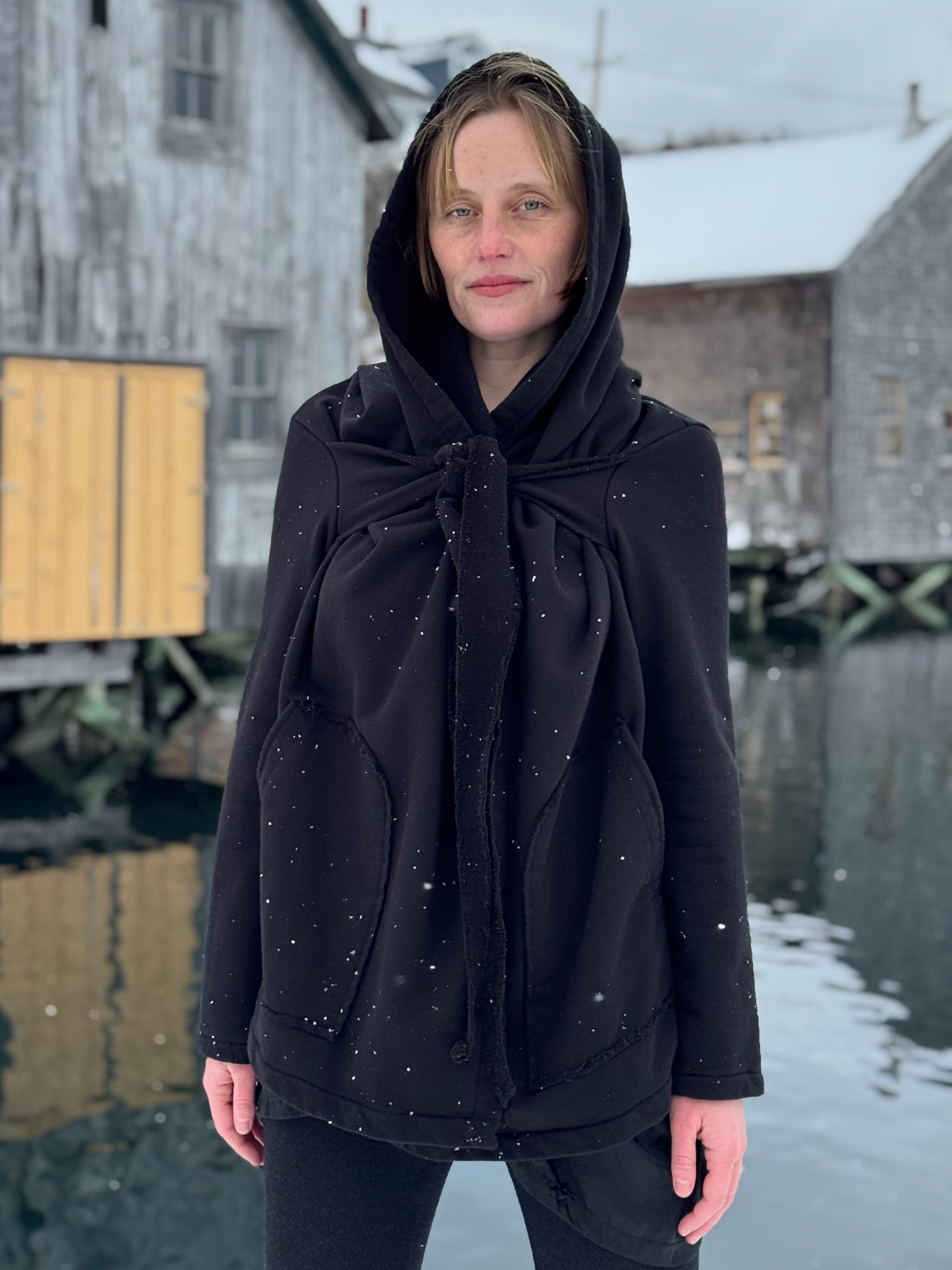 Person wearing a black hooded jacket standing by a waterfront with buildings in the background.