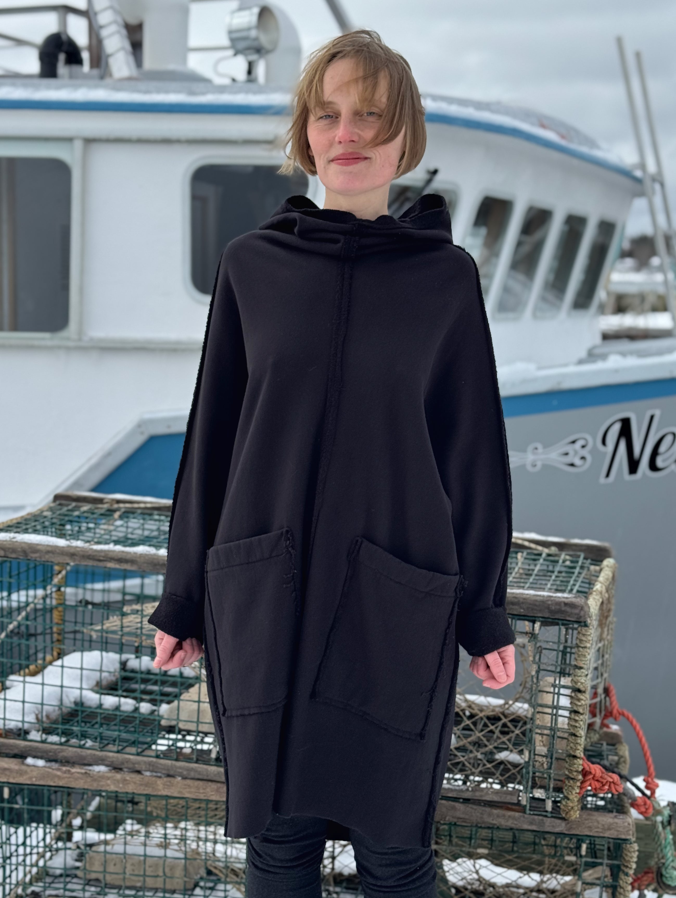Person wearing a black hooded coat standing in front of a boat with lobster traps.