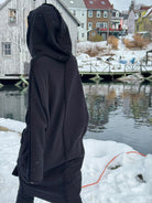 Person wearing a black hooded coat standing by a snowy waterfront with houses in the background.