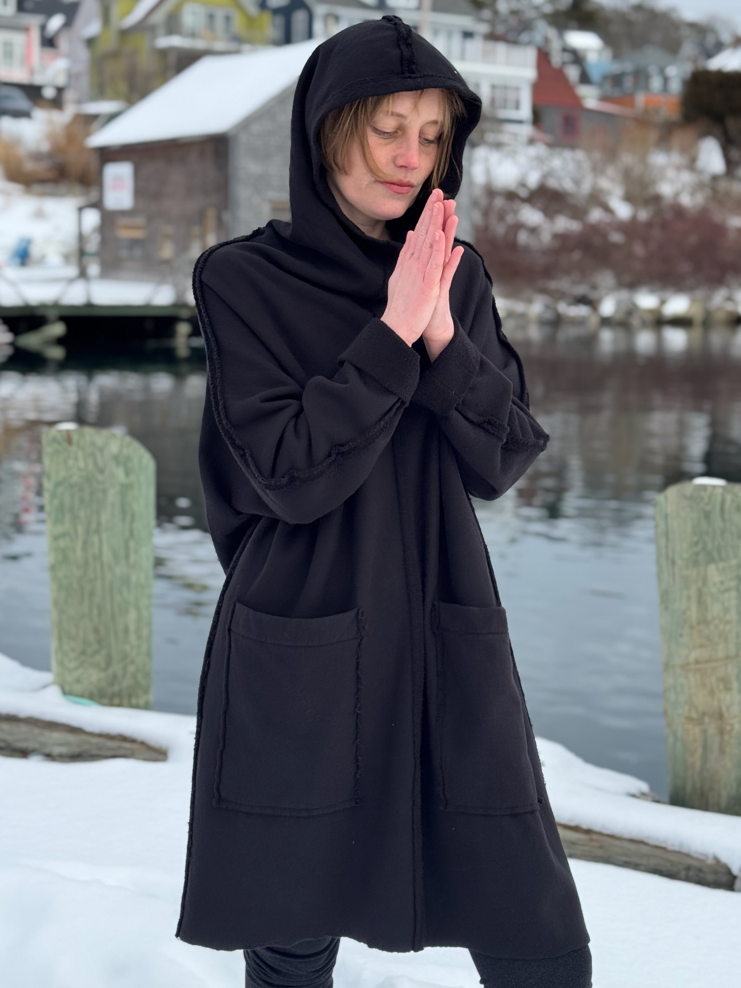 Person wearing a black hooded coat in a snowy landscape with water and buildings in the background.