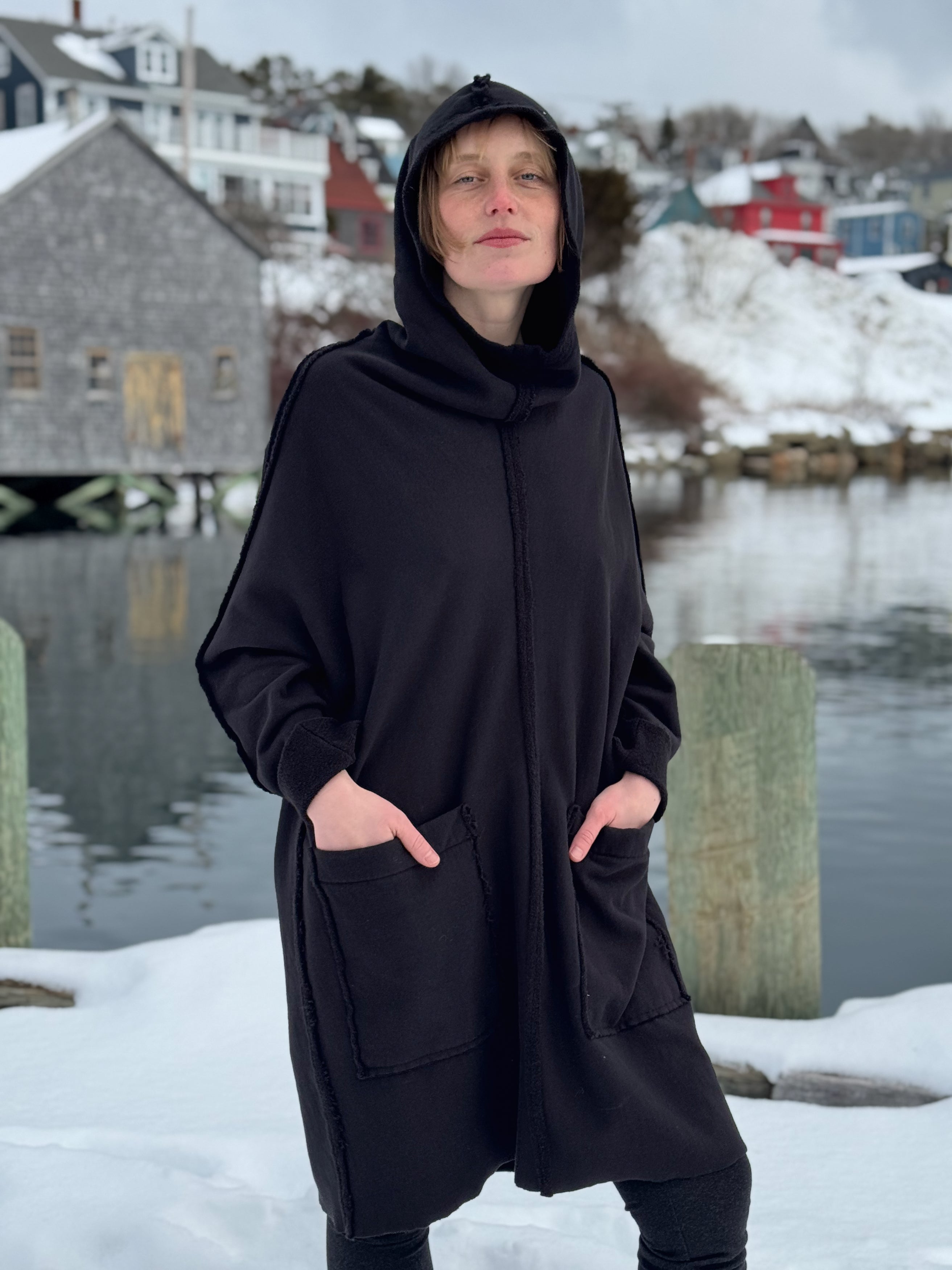 Person wearing a black hooded coat standing in a snowy landscape with water and buildings in the background.