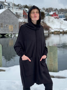 Person wearing a black hooded coat standing in a snowy landscape with water and buildings in the background.