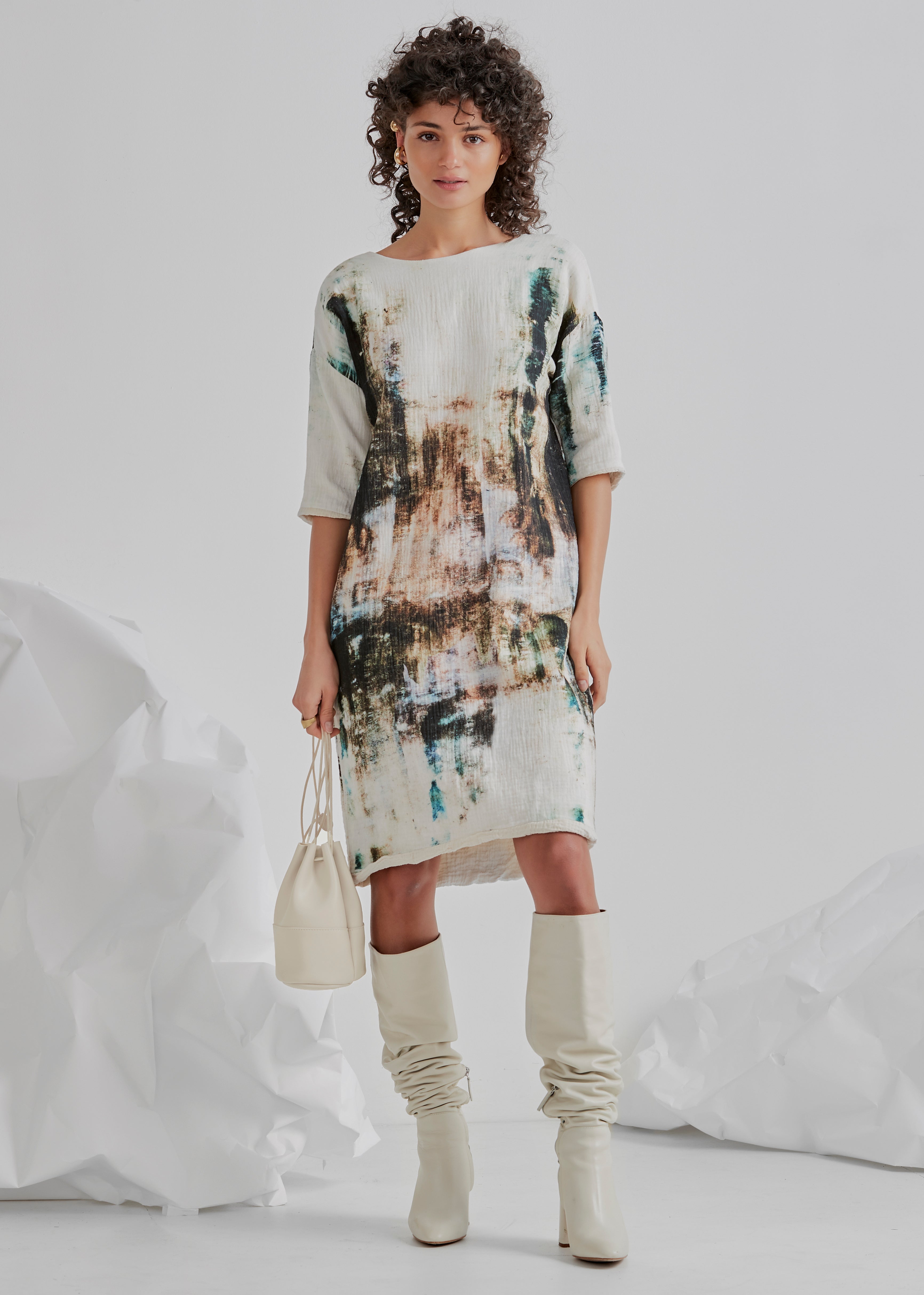 A woman standing and posing in a studio setting, wearing a white and blue abstract print dress with a high neckline and long sleeves, paired with white boots and a beige handbag.